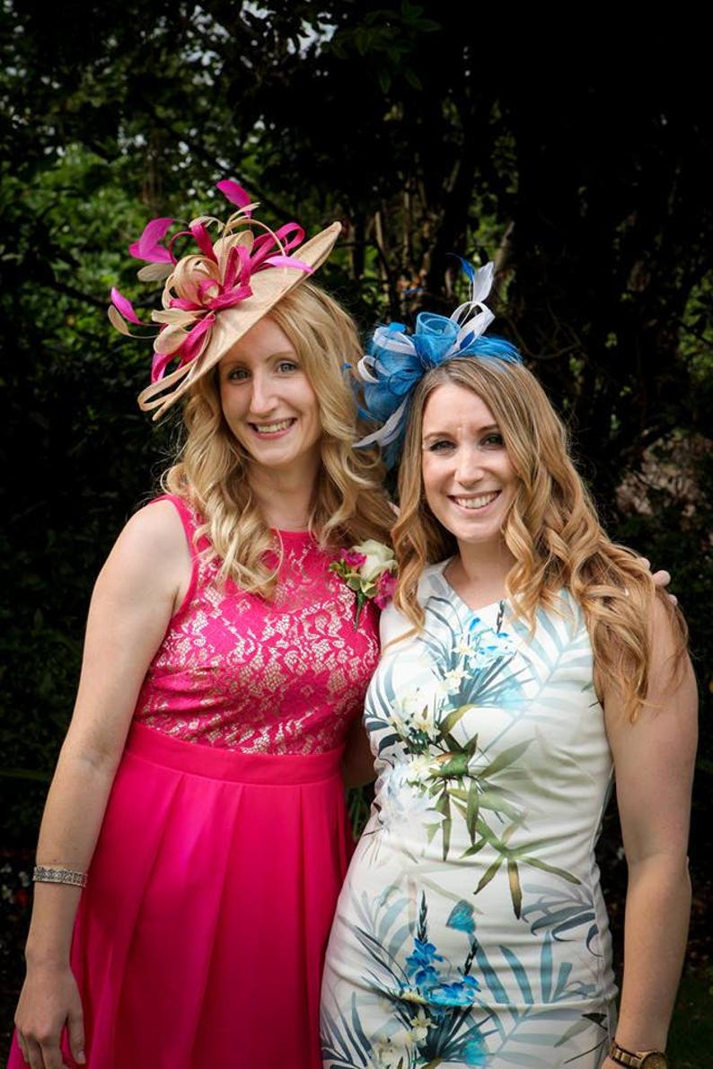 May include: Two women wearing formal attire. One wears a pink dress with a lace bodice and a beige hat with pink and beige accents. The other wears a white dress with a floral print and a blue headpiece.