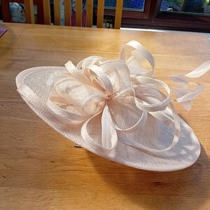 May include: A light peach-colored fascinator hat with a wide brim and decorative bow. The hat features delicate feathers and is suitable for special occasions such as weddings or formal events.