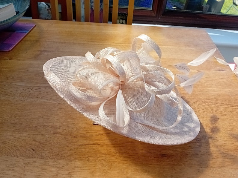 May include: A light peach-colored fascinator hat with a wide brim and decorative bow. The hat features delicate feathers and is suitable for special occasions such as weddings or formal events.
