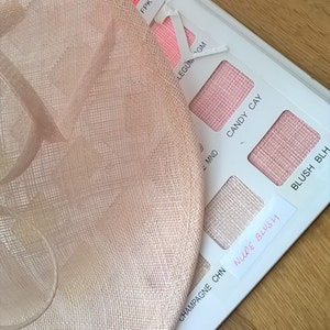 May include: A light peach-colored hat with a wide brim, next to a color swatch card. The card displays various shades of pink and peach, with the handwritten label "NUDE BLUSH". The hat has a woven texture.