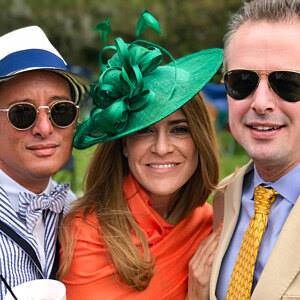 May include: Three people smiling at the camera. A woman in the center wears a bright green hat with a large floral design and an orange scarf. The man on the left wears a white hat with a blue band and a bow tie. The man on the right wears sunglasses and a yellow tie.