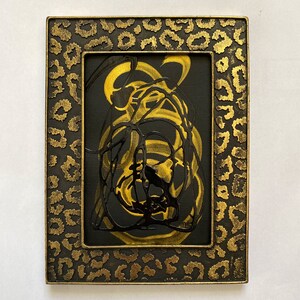 Puede incluir: Obra de arte abstracta en un marco rectangular. La obra presenta un fondo negro con remolinos de pintura dorada y negra. El marco es negro con un diseño de estampado de leopardo dorado.