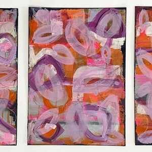 May include: Three abstract paintings in a set. Each painting features a colorful, layered design with pink, purple, orange, and white paint. The paintings are on canvas and have a textured surface.