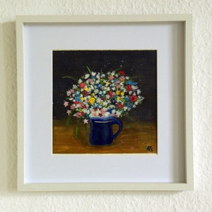 Puede incluir: Pintura enmarcada de un ramo de flores coloridas en una taza azul. La obra presenta un fondo oscuro y una mesa de madera. El marco es blanco.