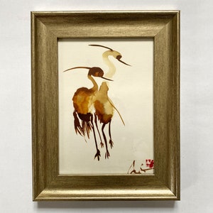Puede incluir: Una pintura de acuarela enmarcada de dos grullas marrones con cuellos y patas largos, sobre un fondo blanco.