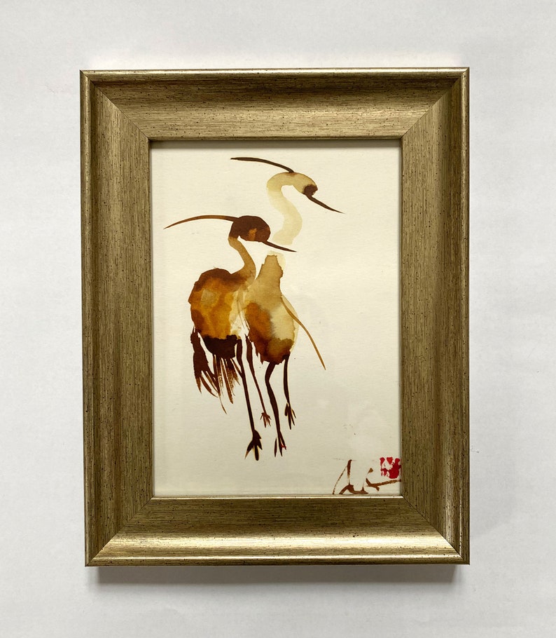 Puede incluir: Una pintura de acuarela enmarcada de dos grullas marrones con cuellos y patas largos, sobre un fondo blanco.