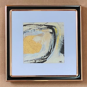 Puede incluir: Obra de arte abstracta enmarcada en un marco negro y oro rosa. La obra presenta un área dorada texturizada con pinceladas negras, blancas y amarillas. La pieza está firmada en la esquina inferior derecha. La obra está montada sobre un passe-partout azul claro.