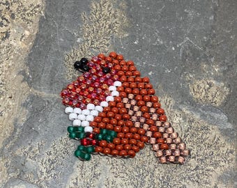 Puede incluir: Adorno de pájaro con cuentas en tonos marrón, rojo, blanco y verde. El pájaro tiene un cuerpo redondeado y un pico pequeño. Las cuentas están dispuestas para crear una superficie texturizada. El adorno es probablemente hecho a mano.
