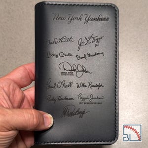 May include: A black leather passport holder featuring the New York Yankees logo and engraved signatures of various baseball players including Babe Ruth, Joe DiMaggio, Mickey Mantle, Darryl Strawberry, Derek Jeter, Paul O'Neill, and Reggie Jackson.