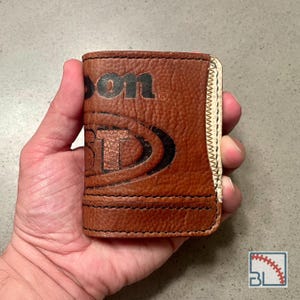 May include: Brown leather wallet with the Wilson logo and a baseball design. The wallet is rectangular, with visible stitching and a white edge. The wallet is held in a hand.