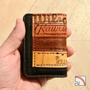 Peut inclure: Un portefeuille rectangulaire en cuir fabriqué à partir de matériaux de gants de baseball recyclés. Le portefeuille présente des panneaux horizontaux dans des tons de beige, marron et noir, avec des coutures visibles et du texte embossé.