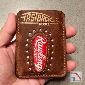 Peut inclure: Un porte-cartes en cuir marron avec le texte FASTBACK MODEL embossé en doré. Le centre présente une découpe ovale révélant un insert en cuir de baseball Rawlings rouge et blanc avec des coutures décoratives autour du périmètre.
