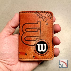 Puede incluir: Una cartera de cuero color canela hecha de una pelota de béisbol, con el texto WILSON POCKET en relieve y un parche circular negro con un logotipo W blanco bordado. La cartera tiene costuras negras visibles a lo largo de los bordes.