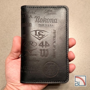 May include: A black leather wallet featuring embossed logos and text including Nokona, Rawlings, Wilson, and Made in U.S.A. The surface displays various baseball-related brand marks and lettering, with visible stitching along the edges of the wallet.