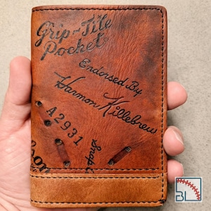 May include: A brown leather wallet featuring the embossed text Grip-Tite Pocket Endorsed By Harmon Killebrew, the number A2931, and the word Snap. The wallet has visible stitching along the edges and a textured surface with dark brown markings.