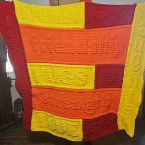 May include: A handmade crochet blanket in shades of red, orange, and yellow. The blanket features the words "FRIENDSHIP", "HUGS", "STRENGTH", and "LOVE" in raised letters. The blanket is approximately 60 inches by 70 inches.