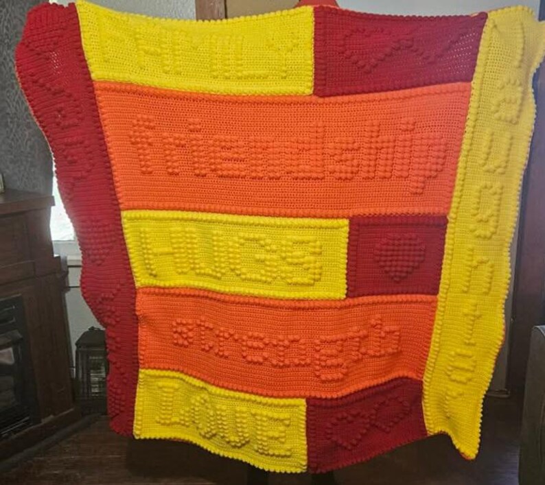 May include: A handmade crochet blanket in shades of red, orange, and yellow. The blanket features the words "FRIENDSHIP", "HUGS", "STRENGTH", and "LOVE" in raised letters. The blanket is approximately 60 inches by 70 inches.