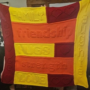 May include: A handmade, colorful crochet blanket with the words "family", "friendship", "hugs", "strength", and "love" stitched into the design. The blanket features a patchwork design with yellow, orange, and red blocks, and heart shapes.