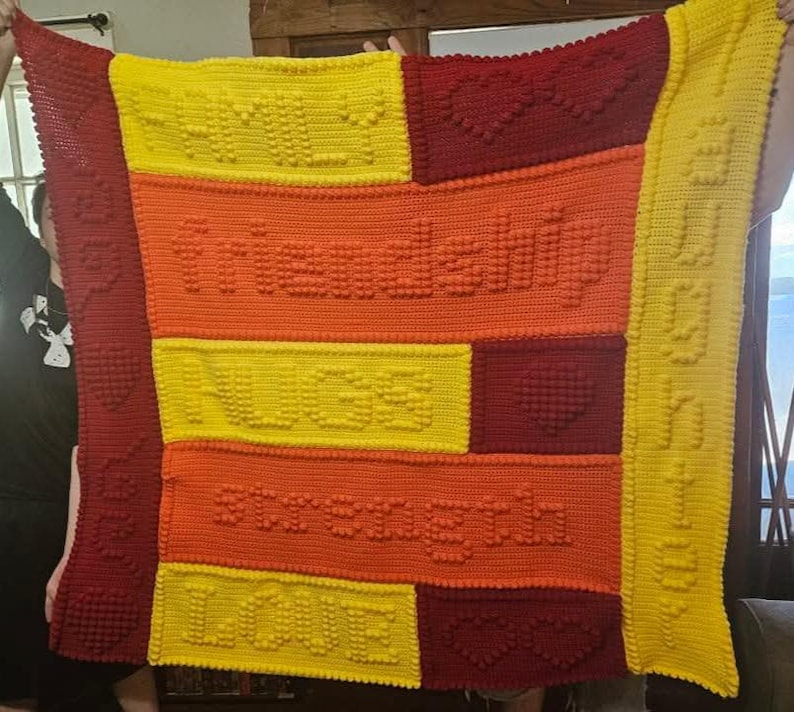 May include: A handmade, colorful crochet blanket with the words "family", "friendship", "hugs", "strength", and "love" stitched into the design. The blanket features a patchwork design with yellow, orange, and red blocks, and heart shapes.