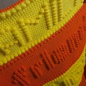 May include: Handmade knitted blanket in yellow and orange. The blanket features a textured pattern with raised bobbles and a central orange stripe with the word "Autumn" knitted into the design. The blanket is a cozy, warm accessory.
