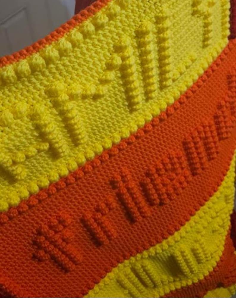 May include: Handmade knitted blanket in yellow and orange. The blanket features a textured pattern with raised bobbles and a central orange stripe with the word "Autumn" knitted into the design. The blanket is a cozy, warm accessory.