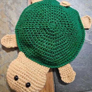 May include: A handmade crochet turtle toy. The turtle has a green shell and a tan head, flippers, and tail. The shell is a dark green color, and the head and limbs are a light tan color. The turtle has two black eyes.