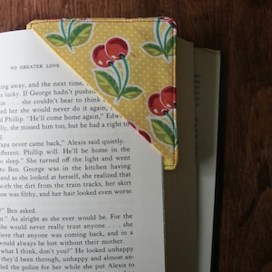 May include: A yellow fabric bookmark with a red and green cherry pattern. The bookmark is folded into a triangle shape and is placed on top of a book. The text "NO GREATER LOVE" is visible on the book.
