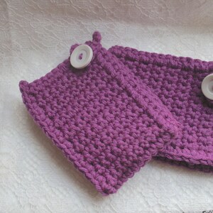 Puede incluir: Un par de calentadores de muñeca de color morado hechos a crochet con botones blancos.
