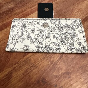 May include: A white floral print wallet with black outlines and a magnetic closure. The wallet is rectangular and features a detailed flower pattern. A small, black magnetic clasp is visible.