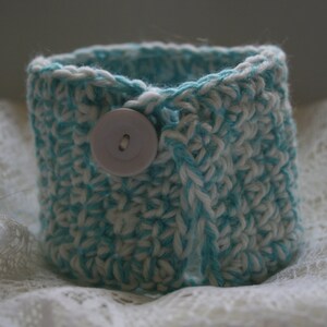 Puede incluir: Un brazalete de crochet en blanco y azul turquesa con un botón blanco.