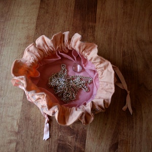 May include: A pink fabric pouch with a ruffled edge, containing a silver chain necklace and a clear glass bead. The pouch is lying on a wooden surface.