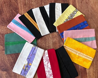 May include: A collection of rectangular fabric pouches in various colors, including pink, green, yellow, and black. Each pouch features a contrasting stripe or design element, such as a patterned fabric or a solid-colored band, adding visual interest to the handmade accessories.