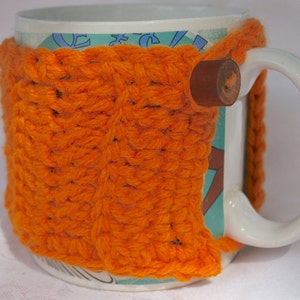 May include: A white ceramic mug with a colorful design and a bright orange crocheted cozy. The cozy has a wooden button.
