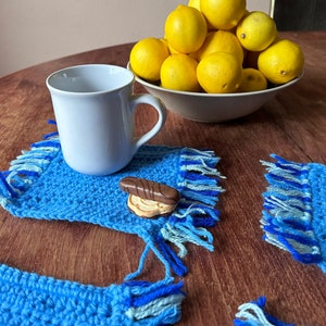 May include: Four blue crocheted coasters with fringe, two with a white mug and a chocolate bar on top, and two empty. The coasters are on a wooden table with a bowl of lemons in the background.