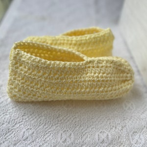 May include: A pair of handmade, light yellow crochet slippers. The slippers are made with a textured stitch, and the open top design allows for easy wear. The soft, pastel color and cozy design suggest comfort and relaxation. The slippers are likely for indoor use.
