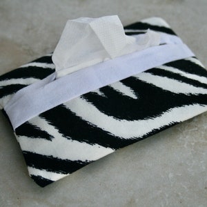 Puede incluir: Una funda para caja de pañuelos de tejido con estampado de cebra en blanco y negro con una solapa de tela blanca.
