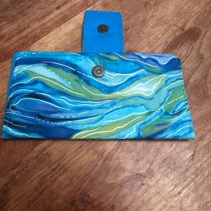 May include: A blue and green patterned wallet with a magnetic snap closure. The wallet features a vibrant, abstract design with shades of blue, green, and white. The flap is a solid blue color.