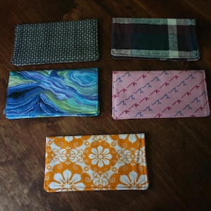 May include: Five fabric pouches with different patterns. The pouches are all the same size and shape. The patterns include a black and white geometric pattern, a blue and green watercolor pattern, a pink pattern with red lines, a plaid pattern, and an orange and white floral pattern.