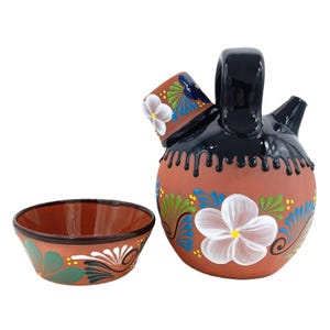 May include: A terracotta pitcher with a blue and white floral design and a matching terracotta cup. The pitcher has a black handle and a black rim.