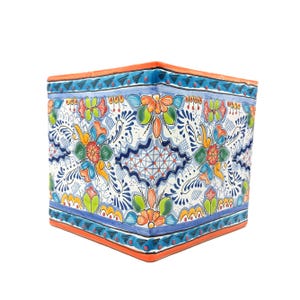 May include: A square ceramic planter featuring a hand-painted floral pattern in vibrant shades of orange, blue, green, and yellow against a white background. The top and bottom edges are trimmed with a solid orange border and blue accents.