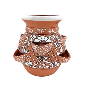 May include: Terracotta strawberry pot with white floral and geometric designs. The pot has multiple side pockets for planting and a white rim. The design includes white dots and lines, creating a decorative and functional garden item.