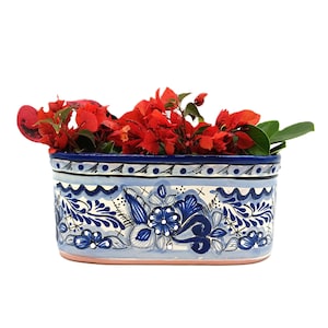 May include: Rectangular ceramic planter with a blue and white floral design, filled with vibrant red flowers and green leaves. The planter has a light pink base and a dark blue rim, showcasing intricate hand-painted details.