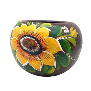 May include: A hand-painted, round, brown ceramic pot. The pot is decorated with a large yellow sunflower with a brown center, green leaves, and white accents. The sunflower design is painted on the front of the pot.