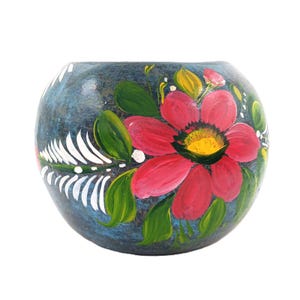 May include: A hand-painted, round ceramic vase with a dark blue background. The vase features a large red flower with a yellow center, green leaves, and white and green accents. The vase is a decorative item.