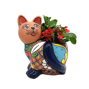 May include: A ceramic cat-shaped planter with a brown body, blue accents, and colorful floral patterns. The planter holds red flowers and green leaves. The cat has blue eyes and a smiling expression.