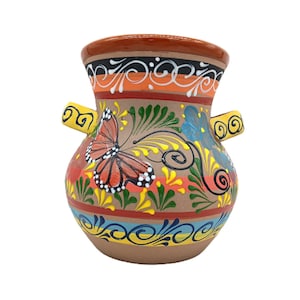 May include: A hand-painted ceramic vase with a brown base and rim, adorned with a vibrant butterfly and floral design. The vase features yellow, orange, blue, and green accents, and two small handles. The overall design is a colorful, decorative piece.