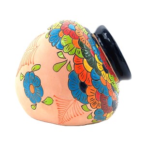 May include: A peach-colored ceramic decorative jar with a black rim. The jar is adorned with a vibrant floral pattern in shades of blue, green, orange, yellow, and red. The design includes stylized flowers and leaf motifs.