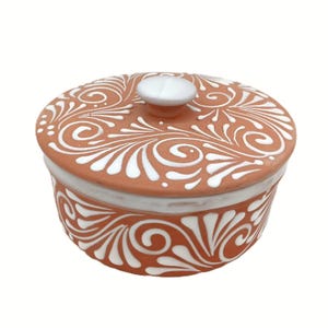 May include: A terracotta-colored ceramic jar with a lid, adorned with white, swirling, floral patterns. The jar has a white rim and a small, white knob on the lid. The design is symmetrical and decorative.
