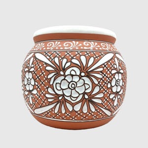 May include: A round terracotta pot featuring a white painted floral pattern and a cross-hatched grid design. The vessel has a smooth white rim and a wide, bulbous body decorated with intricate white flower motifs and thin dark brown outlines.