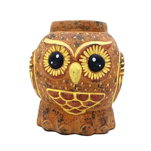 May include: A brown owl-shaped ceramic pot with yellow and black accents. The owl has large, round black eyes, yellow eye details, and a yellow beak. The pot has a textured surface and is likely used for decorative purposes.